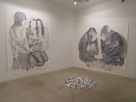Gallery installation view of two large ballpoint pen drawings. On the right is a wall-sized ballpoint pen drawing of three kneeling figures. On the left is a wall-sized ball point pen drawing of two apes facing one another. In front of them are a pile of matchboxes arranged on the floor in a circle. 