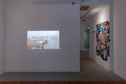 Installation view of two walls in the exhibition. On the left wall, a projection is projected onto a white surface. Leading into the next room, in the hallway, a bright spotlight lights up a collage of photographic images. 