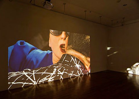 A large projection on a gallery wall shows a person in blue speaking into someone's ear.