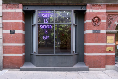 View from the street of a window in the exhibition featuring a purple neon sign that states, "GAY IS GOOD IS GAY."