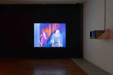 Installation view of a large screen hanging on a dark wall in one of the gallery spaces. The room is dark, but is lit up by the screen. 