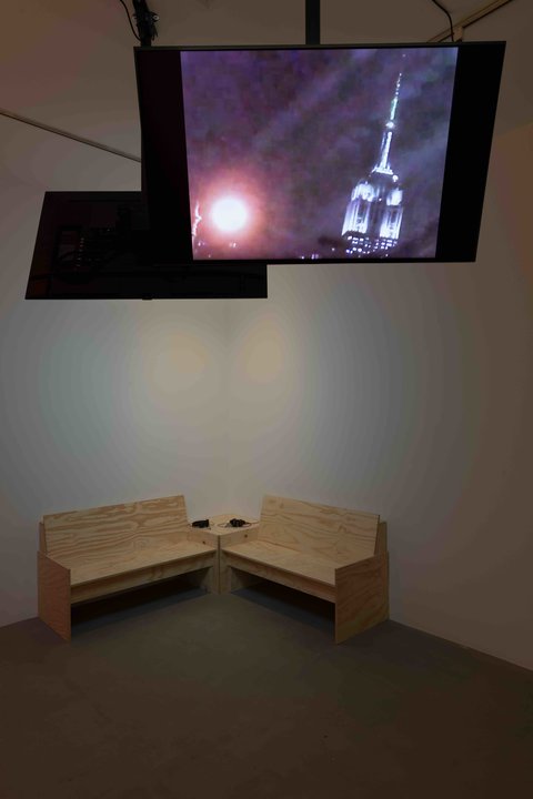Installation view of a room in the exhibition. In the background, two wooden benches are pushed up against the wall and meet at the corner. Two television monitors hang from the ceiling facing away from one another. 