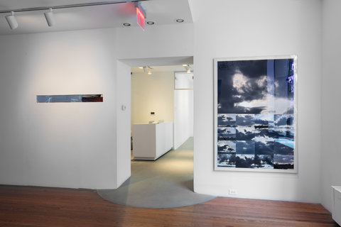 Installation view of two walls in the exhibition separated by a doorway that leads to the lobby of the gallery, where there is a big white desk. On the left, part of the wall is removed to put in a narrow but long mirror. On the right, a photographic image is blown up to be almost the size of the wall. The image is a collage of several images featuring the sun rays peering through clouds. 