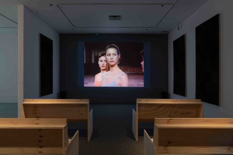 View of the installation of a video in the exhibition. The video is projects against a dark wall at the front of the room. The backs of the wooden benches, arranged like church pews, can be seen from the camera angle at the back of the room. The white walls have large foam pads to absorb the sound. At this moment, the still from the video in the installation view image features to dancers one in front of the other from the torso up as they look off into the distance. 
