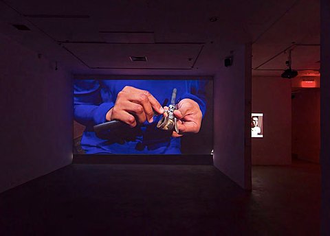 Two large projections can be seen, one in an adjacent gallery room down the hall from the other. The nearer one shows the hands of someone wearing a blue outfit.