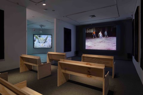 View of the installation of a video in the exhibition. The video is projects against a dark wall at the front of the room. The backs of the wooden benches, arranged like church pews, can be seen from the camera angle at the back of the room. The white walls have large foam pads to absorb the sound. At this moment, the still from the video in the installation view image features a dancer in a blue leotard. In the back of the still, another dancer wears a blue leotard in the shadows. 