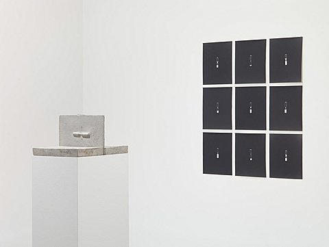 A three-by-three grid of black drawings hang behind a pedestal holding a small white sculpture.