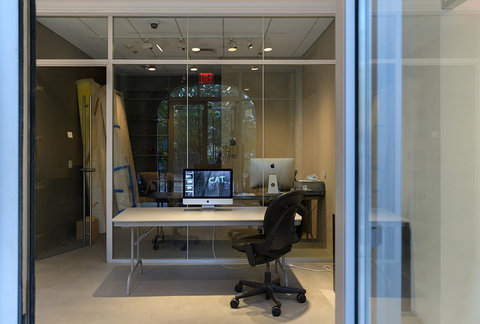 Installation view of the exhibition featuring a black rolling desk chair in front of a grey table. On the table, is an iMac computer with a still from a video on it. Behind the computer, are tall glass windows that separate the first room in the foreground from the room in the background. 
