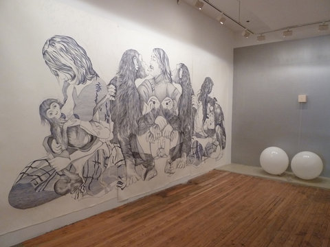 Gallery installation view of two walls. On the left is a floor to ceiling ballpoint pen drawing of many figures interacting with one another. Their bodies are intertwined and engaged. On the left wall, which has been painted light grey, there are two white spherical objects sitting next to each other on the bottom of the wall almost touching the floor. 