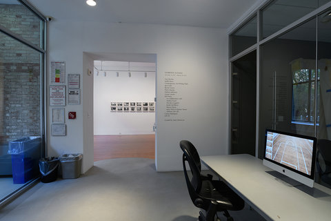 Installation view of the exhibition. On the left, there is a floor to ceiling window. In the middle, a doorway leading to the next room in the exhibition. On the left of the doorway, various posters and notices are hung up in random order, but are illegible from camera view. On the right of the doorway, wall text is displayed, vertically, but again illegible from camera view. On the very right side of the image, a grey table is set up with a black, rolling office chair. On the table, an iMac desktop computer sits with a still image from a video playing on the screen. Behind the computer, more floor to ceiling windows separate one room from the next. 