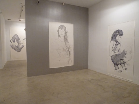 Gallery installation view of two drawings, leading into hallway on the left that gives a sneak preview of the next drawing in the following room. On the left, is a large, portrait of a figure with long hair. Their hands are on their hips and they are looking off to the left. The figure has on a button down top but no pants to reveal their vulva. 