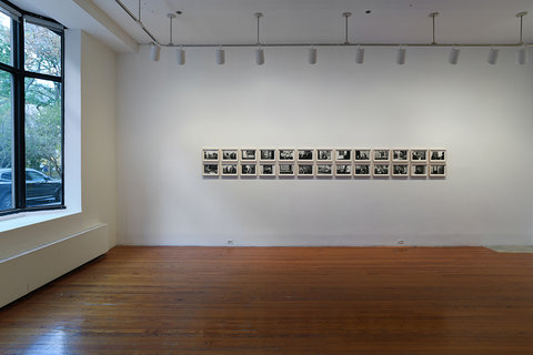 Installation view of the exhibition. On the right, a large window takes up three fourths of the wall. On the back wall, several black and white images are hung in two rows of thirteen. 
