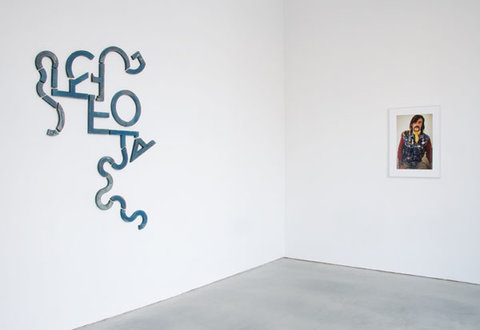 Gallery installation view of left and right wall. On the left is a blue, abstract line sculpture on the wall. On the right is a painting, portrait of a man with a mustache whose gaze stares straight into the viewer. 