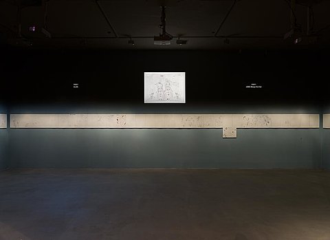 A film is projected on the top half of a darkened gallery wall. A strip of drawings, lighted with direct light, runs along the middle of the wall.