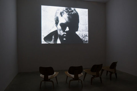 Installation view featuring a still from a video screening. Four chairs sit in front of a black and white image of a mans face in a dark room. 