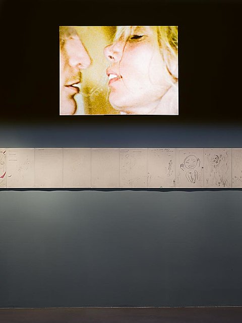 A film is projected on the top half of a darkened gallery wall. A strip of drawings, lighted with direct light, runs along the middle of the wall.