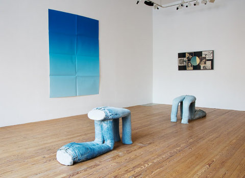 Installation view with four artworks, with two sculptures on the floor, a painting on the left wall, and a painting on the back right wall. The front sculpture features two pairs of jeans, filled with cement. On pair is supporting the other pair with folded legs at the knees. Behind the work, is another jean sculpture. On the left wall is a blue gradient painting. Behind, on the right wall is a collage of old portrait photographs and images.  