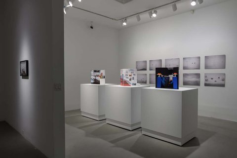 Installation view of three pedestals in the exhibition. On the left side, a white pedestal with a pamphlet on top. In the middle, a white pedestal with another white pamphlet on top and various illegible sketches. On the right, a white pedestal with a black and blue pamphlet on top. In the background, several black and white images are mounted to the wall in two rows of five. In the foreground, a black and white photograph is spotlight in a dark room against a white wall. 