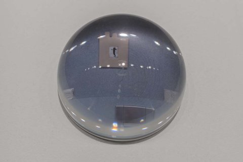 Close-up image of a glass orb in the exhibition atop a white pedestal. The glass orb's convex shape allows for the image underneath to show through in a bit of a distorted way. 