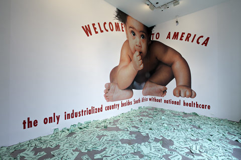 A photo of a giant baby covers the corner of two walls. Behind the baby a text reads: welcome to America, the only industrialized country besides South Africa without national healthcare. Dollar bills are scattered on the floor.