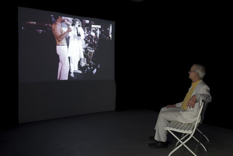 A man in a white suit sits in a white chair facing a projection on a wall in a dark room. The projection features two people on stage playing instruments.