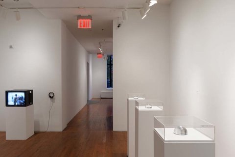 Installation view of the exhibition. On the left, a monitor sits atop a white pedestal. On the right, three white pedestals are lined in a row. 