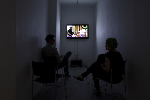 Two people are seated in a dark room facing a television screen that is mounted on the wall. On the screen are three figures. The television is the only source of light in the room. 