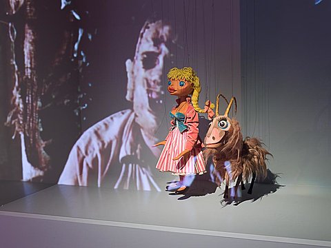 A projection of a masked horror movie character is cast on top of two small marionette figurines.