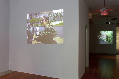 A projection on a white wall. The projection features a woman and the words "Studio of the Streets" in yellow writing over the image. In the background, a darker room features a different projection. In this projection a woman in a green winter coat faces a figure whose back is to the camera. 