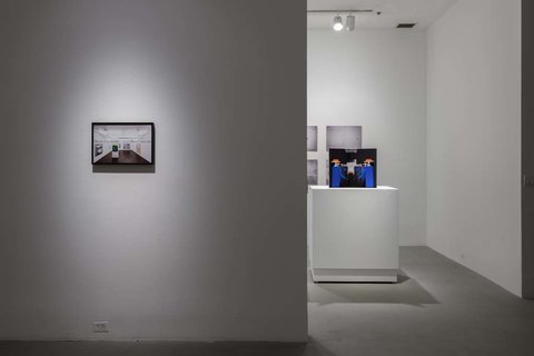 Installation image of the exhibition. On the left, in the foreground, a black and white photograph is mounted to a white wall. A spotlight, lights the image. Leading into the background, a dark colored pamphlet sits atop a white pedestal, but it is illegible from this camera distance. 