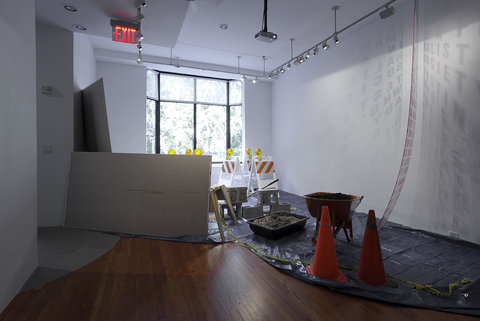 A large window illuminates the room. Around the floor are placed construction objects such as orange cones, a wheelbarrow, dirt, cinderblocks, and drywall on a blue tarp.