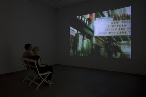 Two people in a dark room are sitting in foldable chairs face a projection on the wall. In the projection there is a blurry projection of outside a movie theater with passers by in the foreground. 