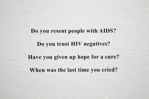 Small black text on a wall that reads the phrases: "do you resent people with AIDS? Do you trust HIV negatives? Have you given up hope for a cure? When was the last time you cried?"