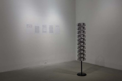 Installation view of the exhibition featuring; on the left wall, five piece of paper with black typed words, though illegible from the camera viewpoint. On the right, near the center of an image, a post card carousel stands with the same image card in each slot. 