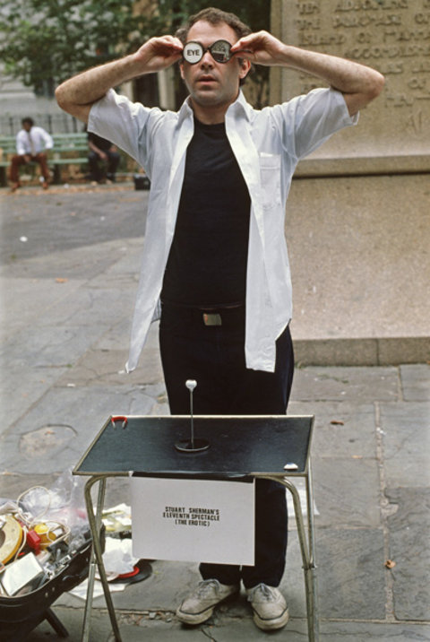 Image of a figure holding his hands up to his head, with his elbows out, he puts glasses on his face. The glasses say "eye" on the left lens. In front of him is a short table with a paper sign hanging from it that states, "Stuart Sherman, Eleventh Exhibition." The figure is wearing a white, short sleeve button down shirt and a black undershirt and black pants.  