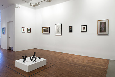 Framed artworks hang on a gallery's walls. A short pedestal on the ground has several sculptures of birds with human faces.