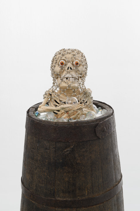 Close-up installation view of a sculpture in the exhibition featuring a brown barrel on which sits the top half of a fake skeleton figure. The skeleton is surrounded by small orbs and marbles and holds a clear orb in its bony hand. It's eyes are wide open and looks up at the viewer with big orange eyes. It is draped with a net of metal chain with small white beads. 