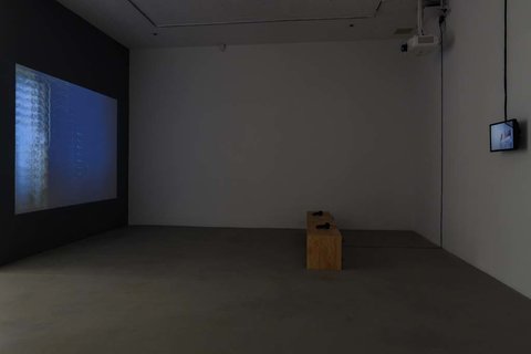 Installation view of the exhibition featuring two walls. On the left, a large screen projects onto a wall. On the right wall, a small television monitor is mounted to the wall. In the middle, a light brown bench sits on the grey floor. The lights are off in the room with only the glow of the televisions and monitors lighting the room. 
