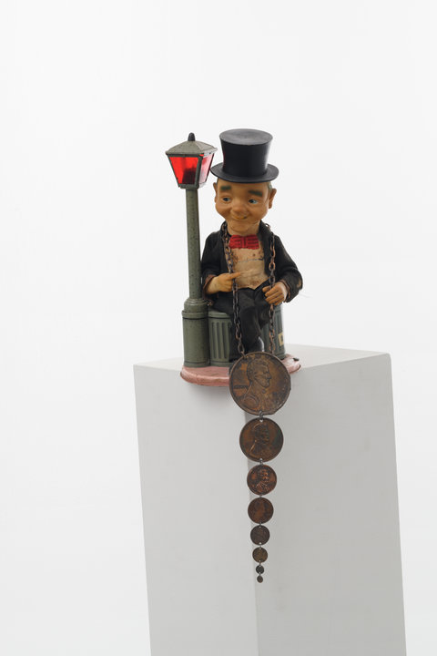 Atop a white pedestal, sits a figurine of a lamppost and garbage can. On the garbage can sits a male figure wearing a black suit, red bowtie, and black top hat. He looks off to the side with a slight smiled expression. A chain is draped around his neck on which hangs eight pennies all descending in size. They hang off the pedestal, below the figurines base. 
