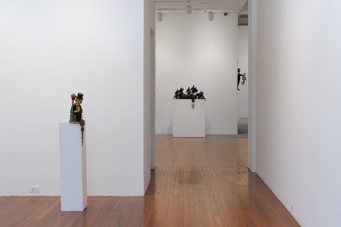 Installation image of two pedestals. Sculptures sit atop the pedestals. One pedestal is against a white wall in the foreground and the other can pedestal sits in the background down a hallway. 