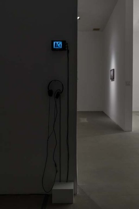 Installation view of a small monitor in the exhibition featuring a small television monitor mounted to the wall. In front of it, a small white box for standing. Also mounted to the wall, a pair of black headphones. 