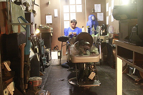 Photograph of a person wearing a bright blue outfit sitting in a cluttered workshop.