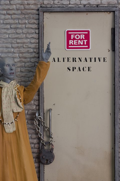 Close-up image of a grey figure wearing a orange robe against a grey brick wall. The figure points at a door in the wall that says, "FOR RENT," on a red sign and below that, "ALTERNATIVE SPACE" in all capital black lettering. The door has a big metal handle, and a is locked with a chain and a padlock. 