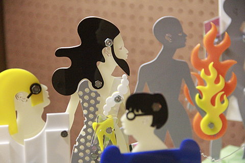 Close up image of multiple flat figurines. The profile of the figure in the center is facing the right, she has fair toned skin and long wavy dark hair. 
