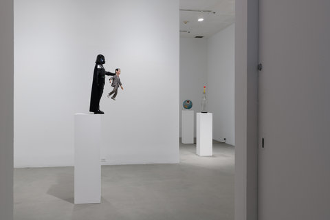 Installation view of the exhibition featuring a Darth Vader character figurine that is dressed in all black holding a smaller figurine by the neck. The figurines are atop a white pedestal. In the background of the image, there is two more pedestals with illegible objects atop them. 