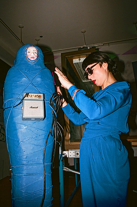 A women with short dark hair, light skin, dark sunglasses and bright red lipstick is wearing a bright blue jumpsuit. She appears to be placing something on a blue sculpture that looks like a mummy. 