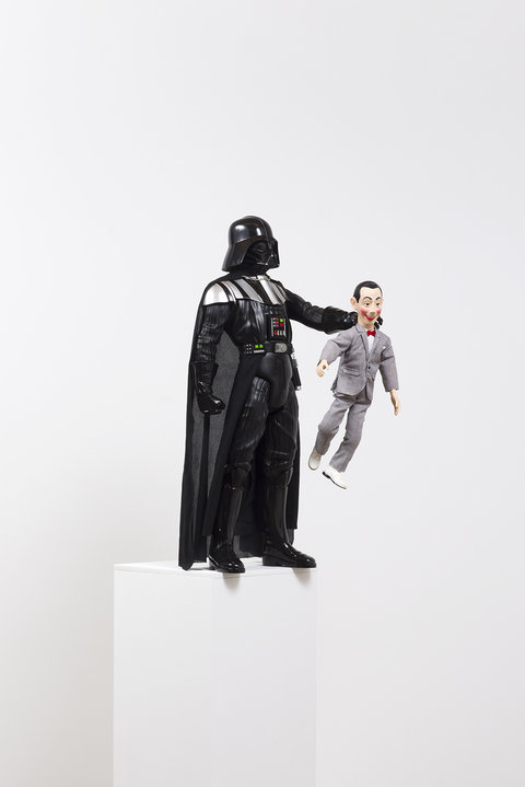 Image of a figurine in the exhibition featuring a Darth Vader character in all black, complete with a helmet and cape, holding the neck of a smaller cheery-eyed figurine wearing a grey suit. The figurine has rosy red cheeks, a big red smile, and a red bowtie. His white choose dangle off of as the Darth Vader character holds him out into the abyss. The figures are displayed on a white pedestal. 