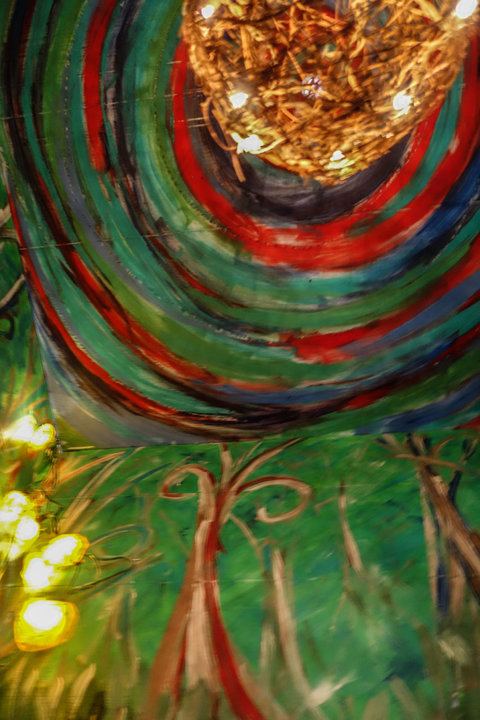 Close-up image of an artwork from the exhibition featuring large portions of green with red, looping stripes near the top of the image. Out of focus, light fixtures made of twine hang in front of the artwork. 