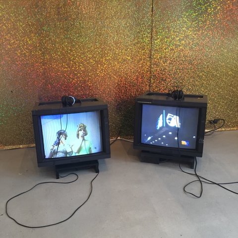 Installation image of the exhibition featuring two TVs on the floor with headphones placed on top of them. On the left, the TV screen features two figures. The figures are shot from below and look down at the camera. They are in front of a light blue background. On the right is a darker TV screen featuring lights that say "54." 