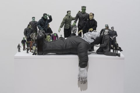 Image of a sculpture in the exhibition featuring several figurines crowded around a larger figurine who seems to have fainted. The figurines include several variations of Frankenstein crowding around a large version of Frankenstein. 